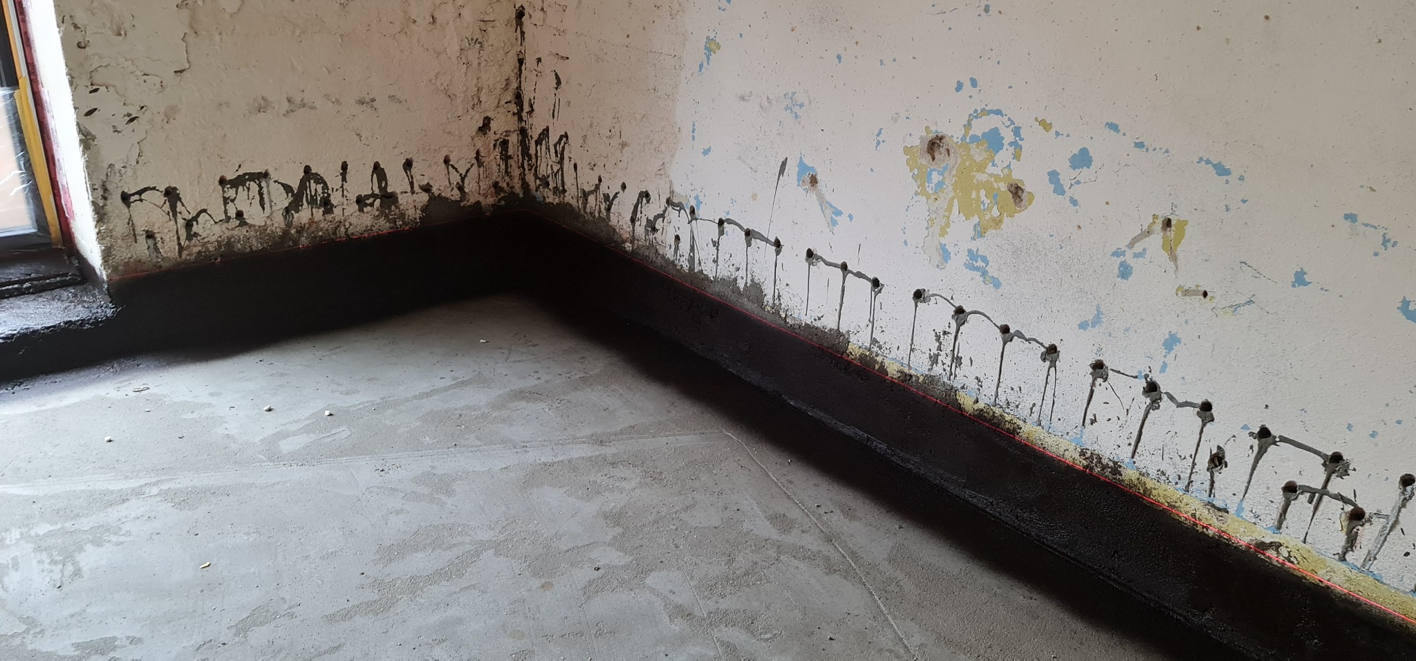 Przygotowanie ściany do remontu: zabezpieczenie dolnej części ściany czarną masą izolacyjną, widoczne nawiercone otwory i ślady po starej farbie.