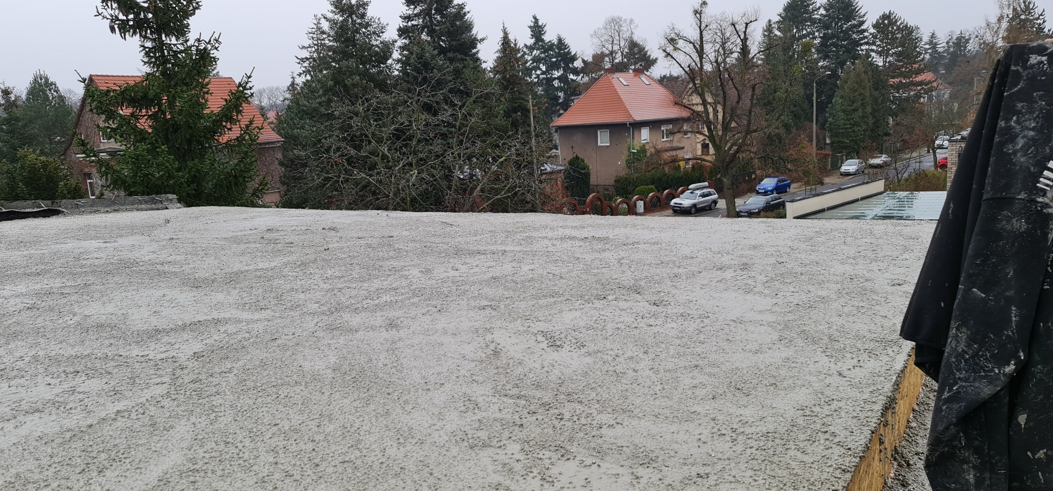 Świeżo wylana, szara wylewka betonowa na dachu budynku w Ostroszowicach, z widokiem na okoliczne domy i drzewa w tle.