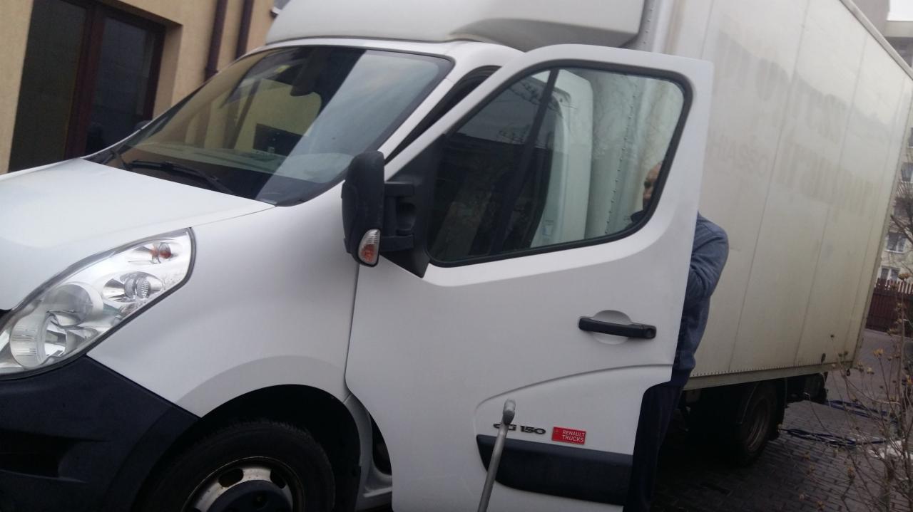 Biały samochód dostawczy Renault Trucks z otwartymi drzwiami kierowcy, widoczny fragment sylwetki osoby w środku, zaparkowany na zewnątrz budynku.