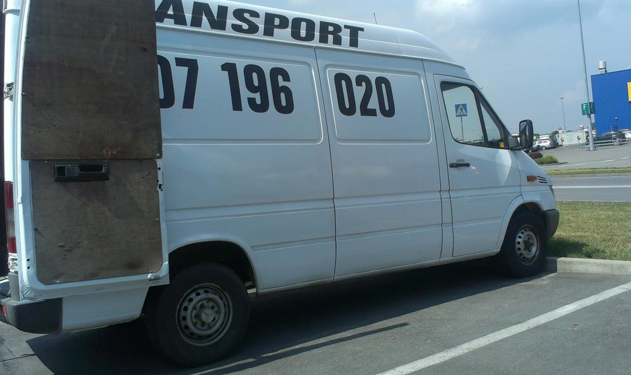 Biały samochód dostawczy z otwartymi, drewnianymi drzwiami z tyłu, zaparkowany na parkingu w słoneczny dzień. Na boku pojazdu duży, czarny numer telefonu.