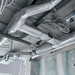 Widok z dołu na system wentylacji i rekuperacji z srebrnymi, zaizolowanymi przewodami w surowym wnętrzu budynku w trakcie wykończenia, widoczne przewody elektryczne i rury.