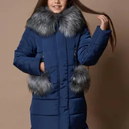 Dziewczynka w granatowej, pikowanej kurtce zimowej z futrzanym kołnierzem i pomponami przy kieszeniach, uśmiechająca się do kamery, na jednolitym beżowym tle.