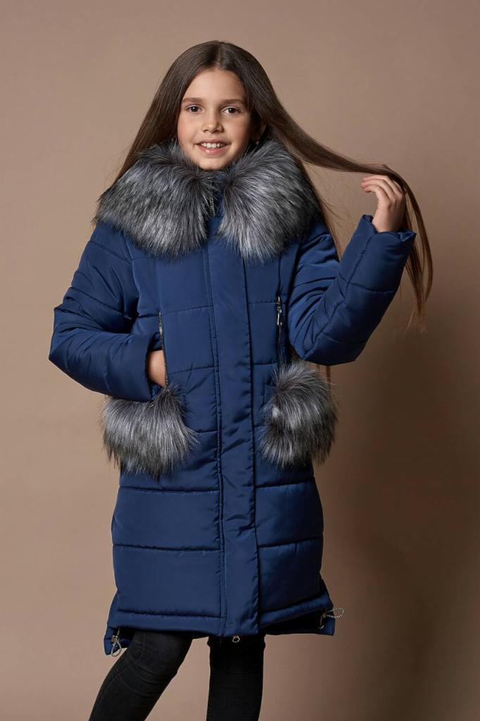 Dziewczynka w granatowej, pikowanej kurtce zimowej z futrzanym kołnierzem i pomponami przy kieszeniach, uśmiechająca się do kamery, na jednolitym beżowym tle.