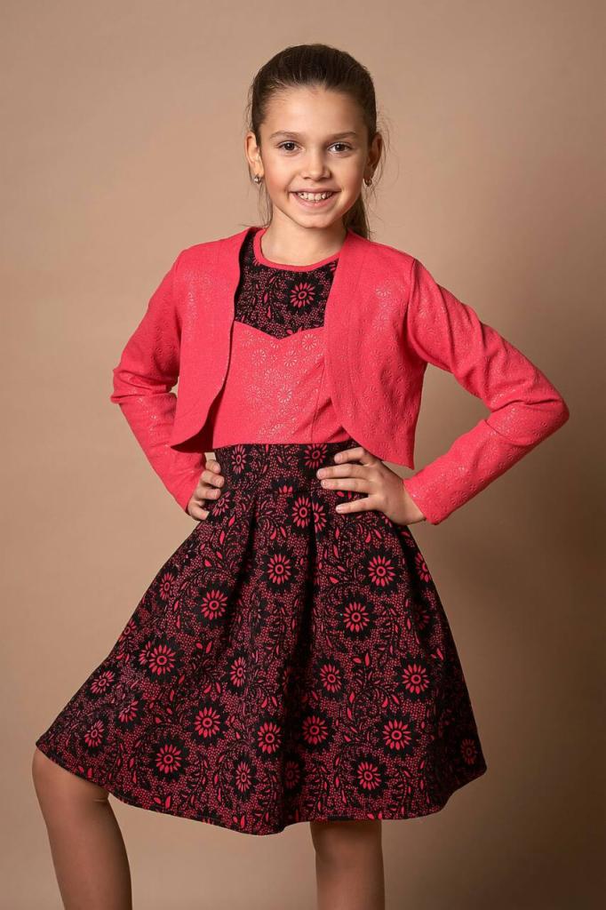 Uśmiechnięta dziewczynka w komplecie: rozpinana, krótka, koralowa narzutka z delikatnym wzorem, koralowa bluzka z czarną koronką przy dekolcie i rozkloszowana spódnica z czarno-koralowym kwiatowym...