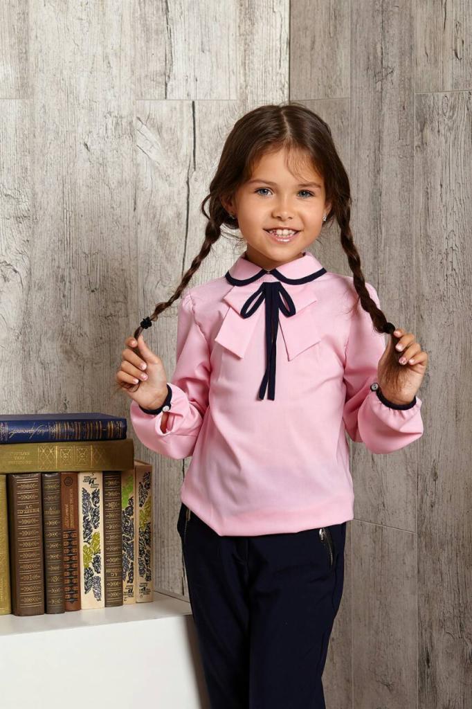 Uśmiechnięta dziewczynka z warkoczami, ubrana w różową bluzkę z granatową kokardą i spodnie, pozuje na tle drewnianej ściany obok stosu starych książek.