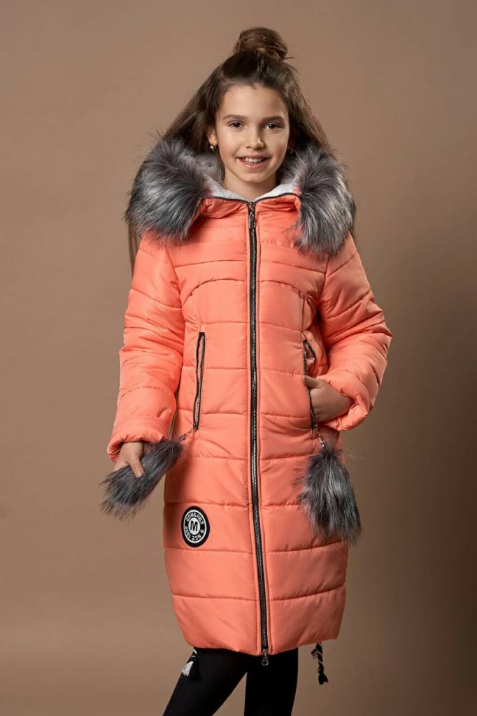 Dziewczynka w długiej, koralowej kurtce puchowej z futrzanym kapturem i pomponami, stojąca na jednolitym tle.