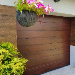 Nowoczesna brama garażowa segmentowa o teksturze drewna w odcieniach brązu, ozdobiona wiszącą donicą z różowymi petuniami i zielonym krzewem po lewej stronie.