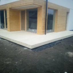 Nowoczesny, surowy taras z jasnego drewna przylegający do budynku z drewnianą elewacją, widoczne czarne podłoże i materiały budowlane.