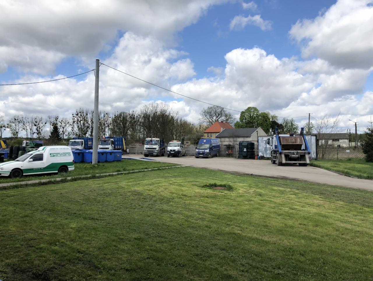 Flota pojazdów specjalistycznych i kontenerów na terenie firmy zajmującej się gospodarką odpadami, widok z perspektywy trawnika.