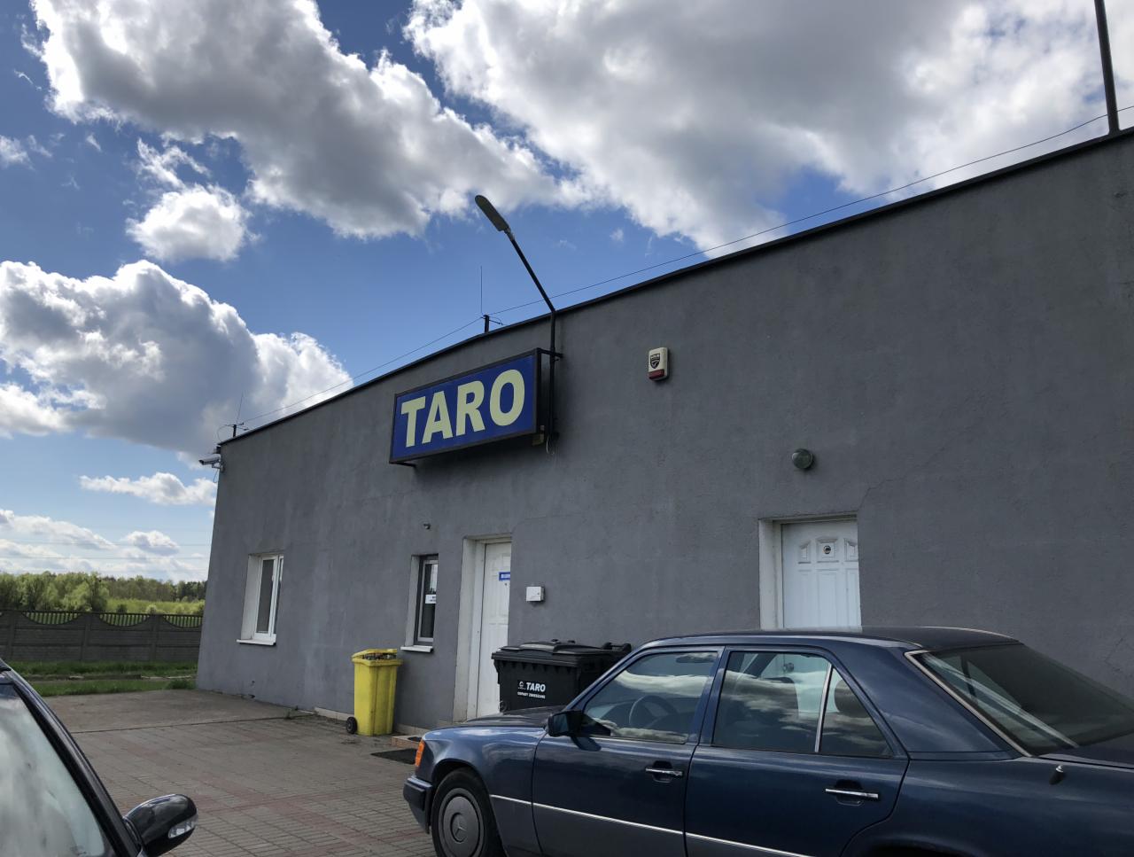 Szary budynek firmy TARO z niebieskim szyldem, zaparkowane samochody osobowe, żółty kosz na śmieci i czarne pojemniki na zewnątrz, błękitne niebo z chmurami w tle.