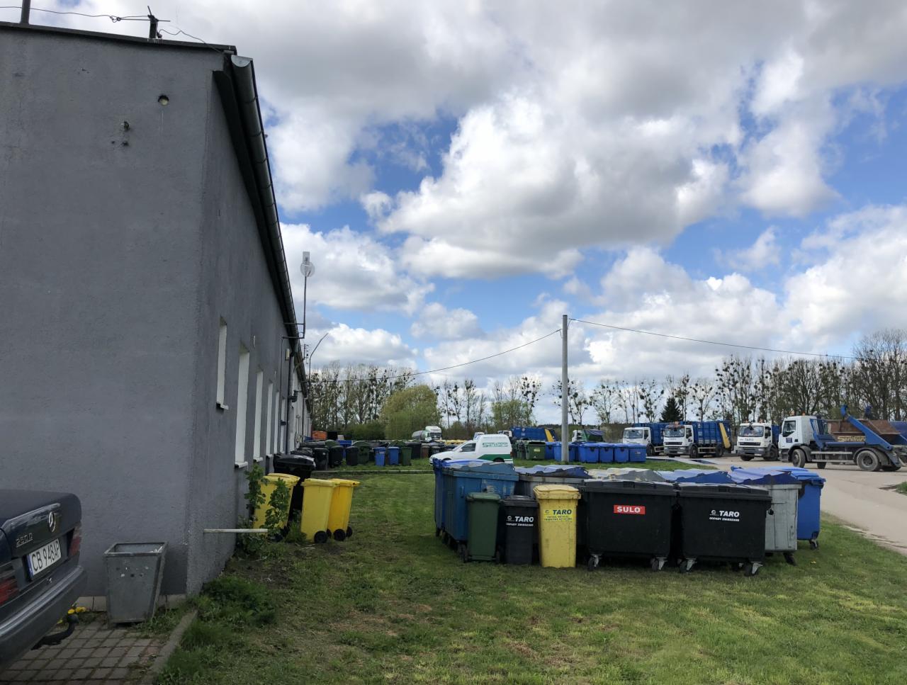 Rząd kontenerów na odpady różnych kolorów (żółte, czarne, niebieskie, zielone) ustawionych na trawie obok szarego budynku, w tle widoczne ciężarówki do wywozu odpadów i drzewa pod błękitnym...