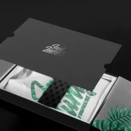 Otwarty czarny box prezentowy z srebrnymi elementami, zawierający białą koszulkę z motywem liści tropikalnych, czarny krawat i dodatkowy materiał, całość na czarnym tle.