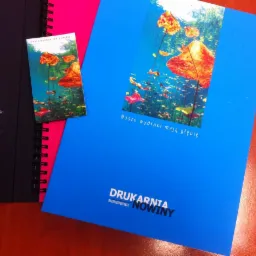 Przykładowe wydruki drukarni: ulotka i katalog z motywem podwodnego krajobrazu z rybami i roślinami, leżące na biurku obok notesu w twardej oprawie.
