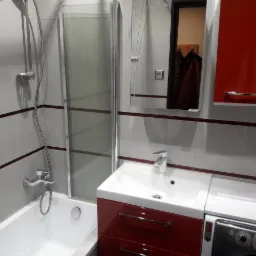 Wykończona łazienka z wanną za szklanym parawanem, umywalką na czerwonej szafce, pralką obok i szafkami wiszącymi w kolorze czerwonym, z płytkami w odcieniach szarości z bordowym paskiem.