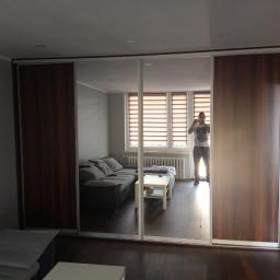 Antracyt Meblarstwo - Szafa przesuwna z lustrami w salonie, odbicie fotografa robiącego zdjęcie, widoczna sofa, okno z roletami i ciemna podłoga.