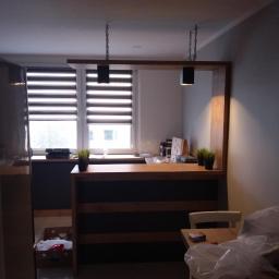 Antracyt Meblarstwo - Drewniana lada z oświetleniem na łańcuchach w nowoczesnym wnętrzu, widok na okno z żaluzjami i fragment stolika z krzesłem przykrytym folią.