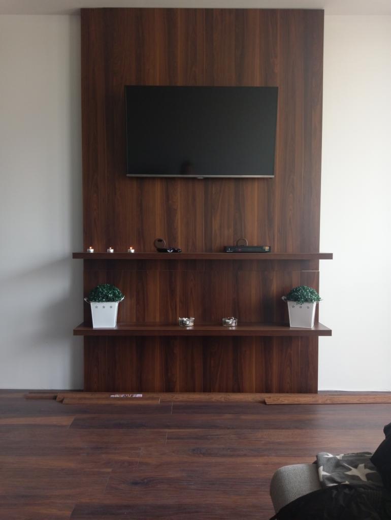Drewniana zabudowa ściany z telewizorem, dwiema półkami, dekoracjami i oświetleniem punktowym, dopasowana kolorystycznie do podłogi.