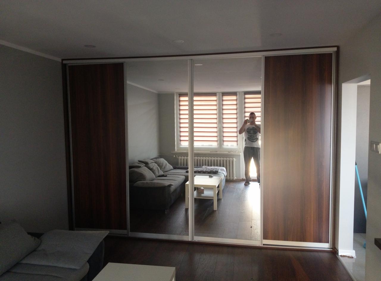 Szafa przesuwna z lustrami w salonie, odbicie fotografa robiącego zdjęcie, widoczna sofa, okno z roletami i ciemna podłoga.