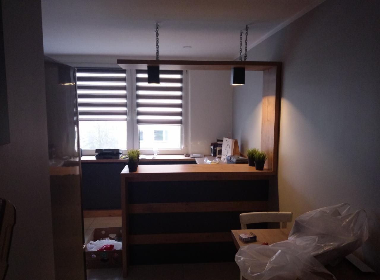 Drewniana lada z oświetleniem na łańcuchach w nowoczesnym wnętrzu, widok na okno z żaluzjami i fragment stolika z krzesłem przykrytym folią.