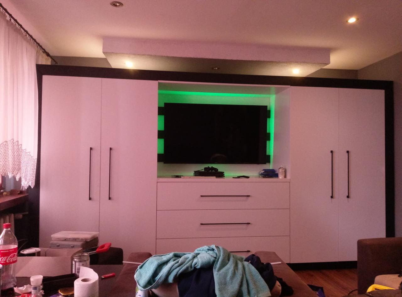 Biała szafa z wbudowanym telewizorem podświetlonym na zielono, widoczne trzy szuflady i długie, czarne uchwyty, na pierwszym planie niedbale rzucony ręcznik.