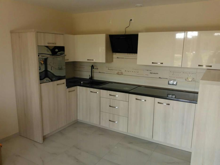 Jasna kuchnia z kremowymi szafkami górnymi na wysoki połysk i dolnymi z drewnopodobnym wzorem, czarnym blatem oraz dekoracyjnym panelem ściennym z motywami kawowymi.