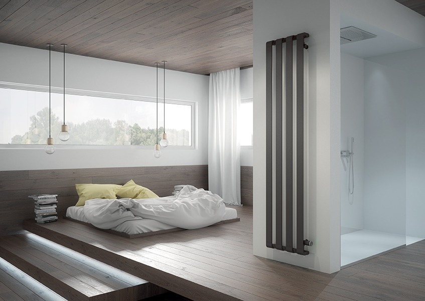 Nowoczesny, pionowy grzejnik w kolorze grafitowym, umieszczony na ścianie między sypialnią z platformą a minimalistyczną kabiną prysznicową.