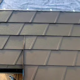 Montaż brązowych dachówek na dachu z widoczną membraną dachową Pro Plus, fragment rynny i elementu zieleni w tle.