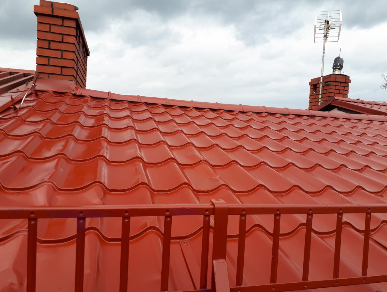 Błyszczący, nowy dach z blachodachówki w odcieniach czerwieni, widok zza pomalowanej na ten sam kolor metalowej balustrady, w tle komin z cegły i pochmurne niebo.