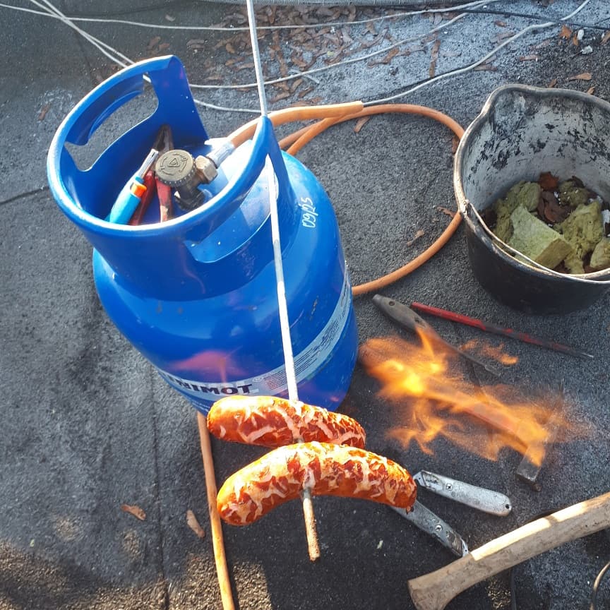 Nietypowe wykorzystanie sprzętu dekarskiego: płonące kiełbaski na dachu obok butli z gazem, wiadra z materiałami izolacyjnymi i narzędzi.