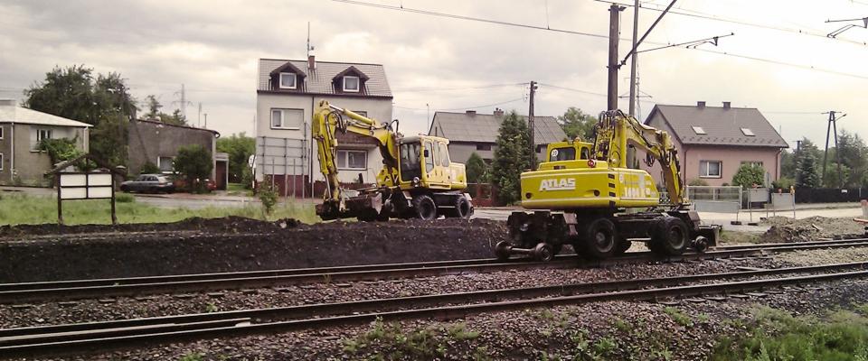 Dwa żółte, kołowe koparki Atlas na torach kolejowych podczas prac ziemnych; w tle budynki mieszkalne i słupy energetyczne.