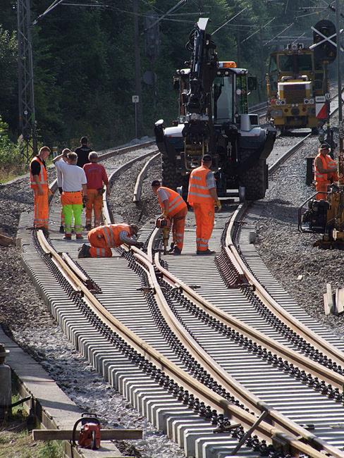 Grupa pracowników w pomarańczowych kombinezonach podczas budowy lub naprawy torów kolejowych; w tle ciężki sprzęt budowlany i linia wysokiego napięcia.