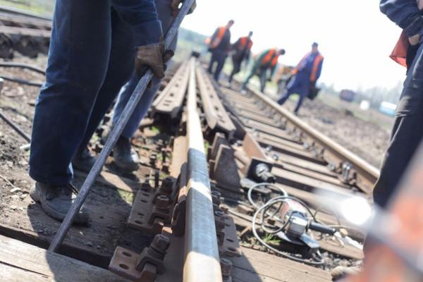 Pracownicy w kamizelkach odblaskowych pracują przy torach kolejowych, używając stalowego pręta i szlifierki kątowej. Widoczne śruby mocujące szyny.