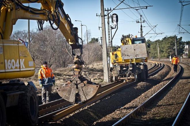 Żółty koparko-ładowarka podnosi metalowe szyny na torach kolejowych, pracownik w pomarańczowej kamizelce obserwuje proces, drugi pracownik idzie po torach w oddali, słupy trakcyjne w tle.