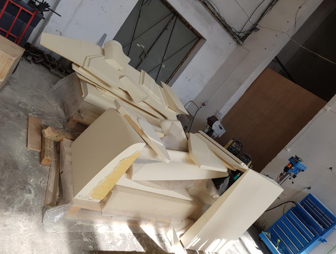 Elementy drewniane, prawdopodobnie do mebli, leżące na palecie w warsztacie, widoczne narzędzia i maszyny stolarskie w tle.