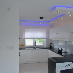 Nowoczesna, minimalistyczna kuchnia z białymi szafkami i czarnym blatem, oświetlona niebieskimi diodami LED w suficie i wyposażona w dotykowy panel sterowania na ścianie.