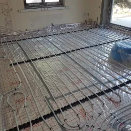 Układanie rur ogrzewania podłogowego na folii z siatką, widoczne czarne uchwyty mocujące i niebieski zwój rury zabezpieczony folią. Surowe ściany i okno w tle.