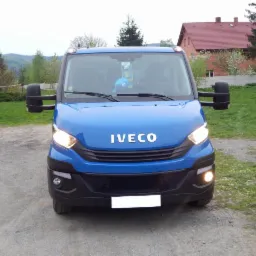 Niebieski bus marki IVECO z włączonymi światłami mijania, stojący na żwirowej drodze z widocznymi drzewami i domem w tle.