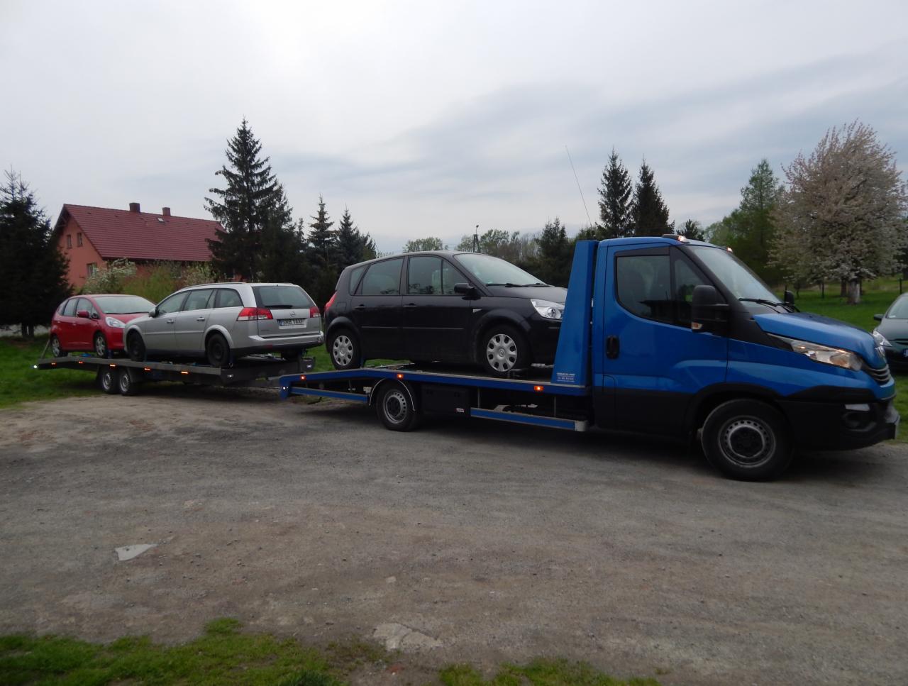 Niebieska laweta z trzema samochodami osobowymi na przyczepie, zaparkowana na żwirowej powierzchni, w tle dom z czerwonym dachem i drzewa.