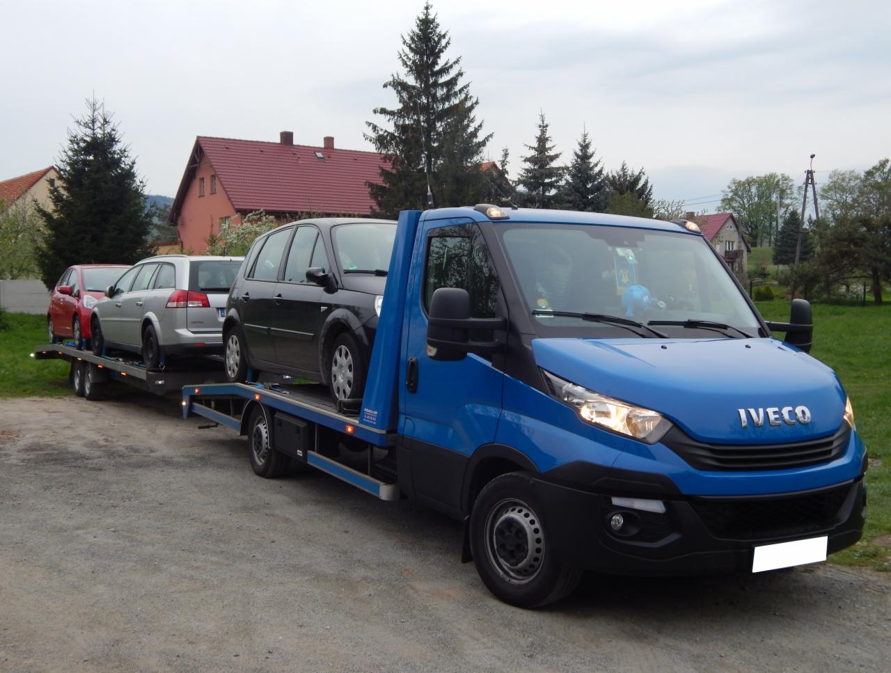 Niebieska laweta Iveco z trzema samochodami osobowymi (czerwony, srebrny, czarny) na platformie, zaparkowana na żwirowej drodze, w tle domy z czerwonymi dachami i drzewa.