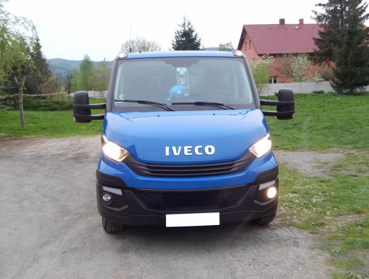 Niebieski bus marki IVECO z włączonymi światłami mijania, stojący na żwirowej drodze z widocznymi drzewami i domem w tle.