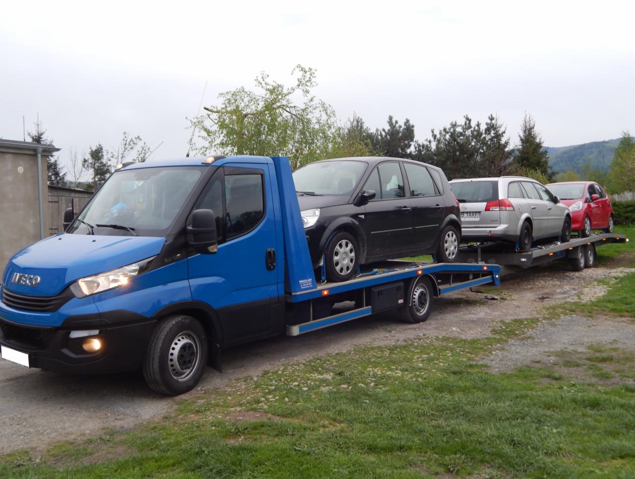 Niebieska laweta z trzema samochodami osobowymi na platformie, zaparkowana na trawiastym terenie z widokiem na drzewa i wzgórza w tle.