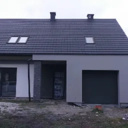 Nowoczesny dom jednorodzinny z szarą elewacją, ciemną dachówką i oknami dachowymi, w trakcie wykończenia terenu wokół budynku. Widoczny stos kostki brukowej i puszki po farbach.