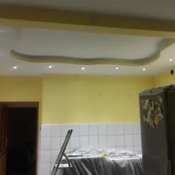 Żółte ściany w kuchni z pomalowanym sufitem z dekoracyjnym wgłębieniem i oświetleniem punktowym, widoczna drabina i zakryty folią blat.