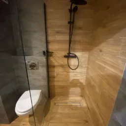 Nowoczesna kabina prysznicowa typu walk-in z deszczownicą w kolorze czarnym, z odpływem liniowym w podłodze wyłożonej płytkami imitującymi drewno, oraz wisząca toaleta na tle szarych płytek.