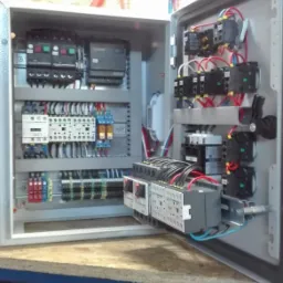 Otwarta szafa sterownicza z widocznymi bezpiecznikami, stycznikami, listwami zaciskowymi i przewodami, zamontowana na drewnianej płycie.