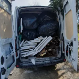 Otwarty bagażnik białego busa wypełniony czarnymi workami i stertą gruzu, w tle drzewo iglaste.