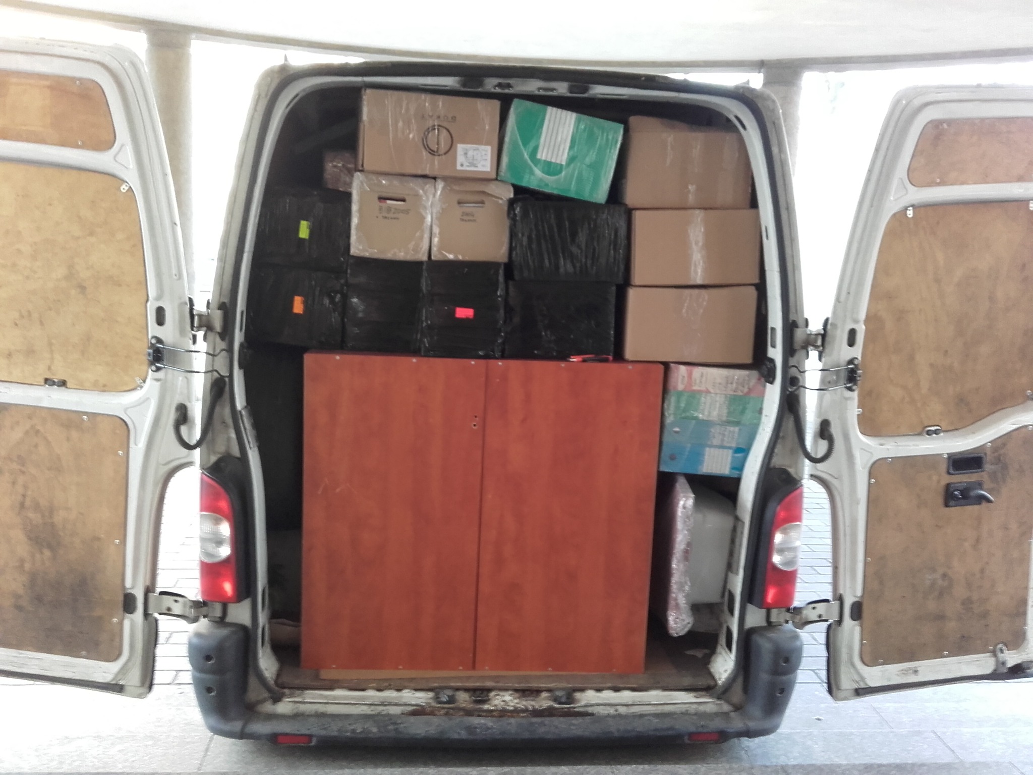 Otwarty bagażnik białego busa wypełniony kartonami, czarnymi workami i drewnianą szafą, widok z tyłu.