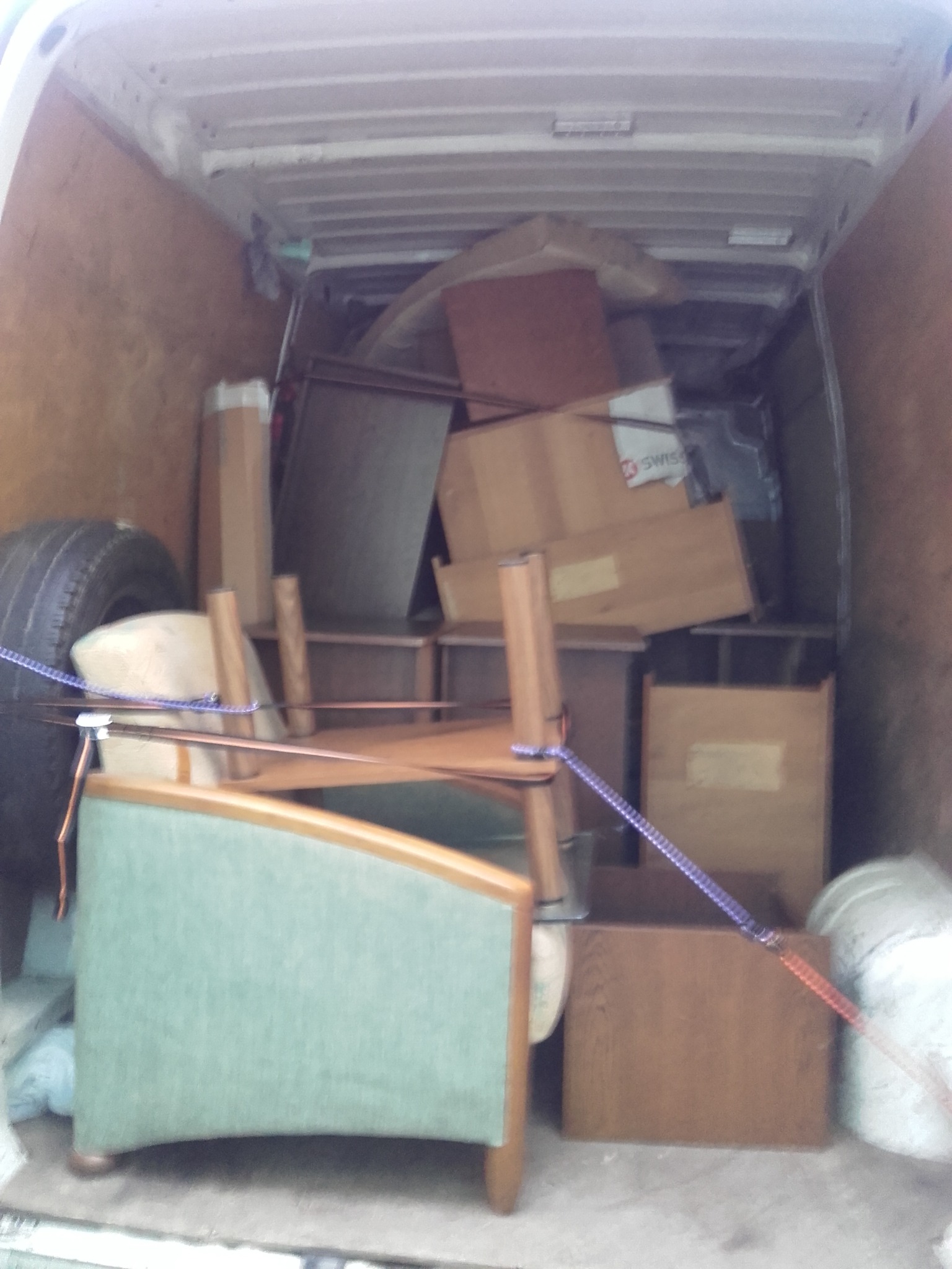 Wnętrze furgonetki załadowane meblami i kartonami, zabezpieczone linami, widoczne opona z boku.