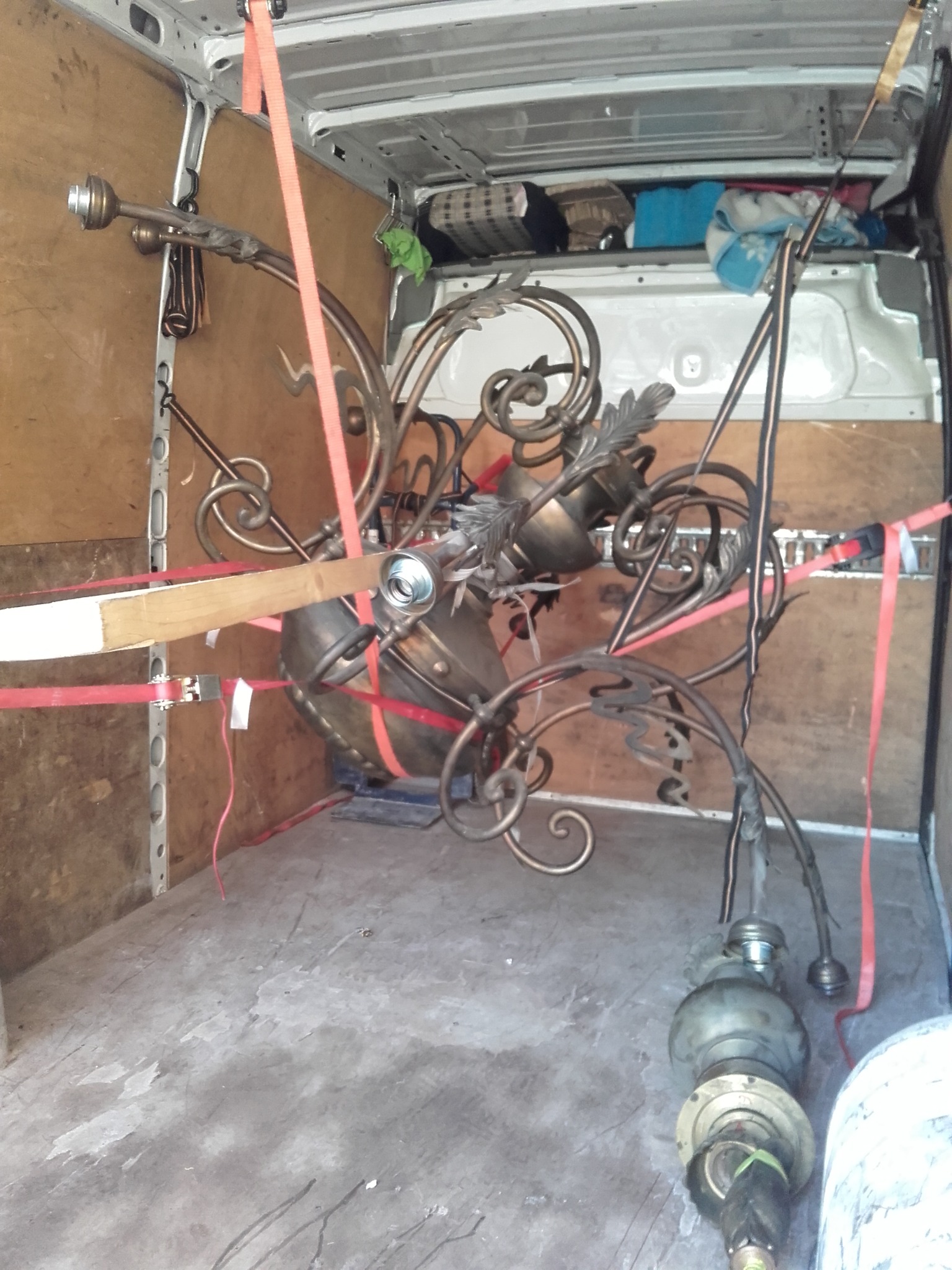 Antyczny żyrandol z metaloplastyki zabezpieczony czerwonymi pasami wewnątrz furgonetki podczas transportu.