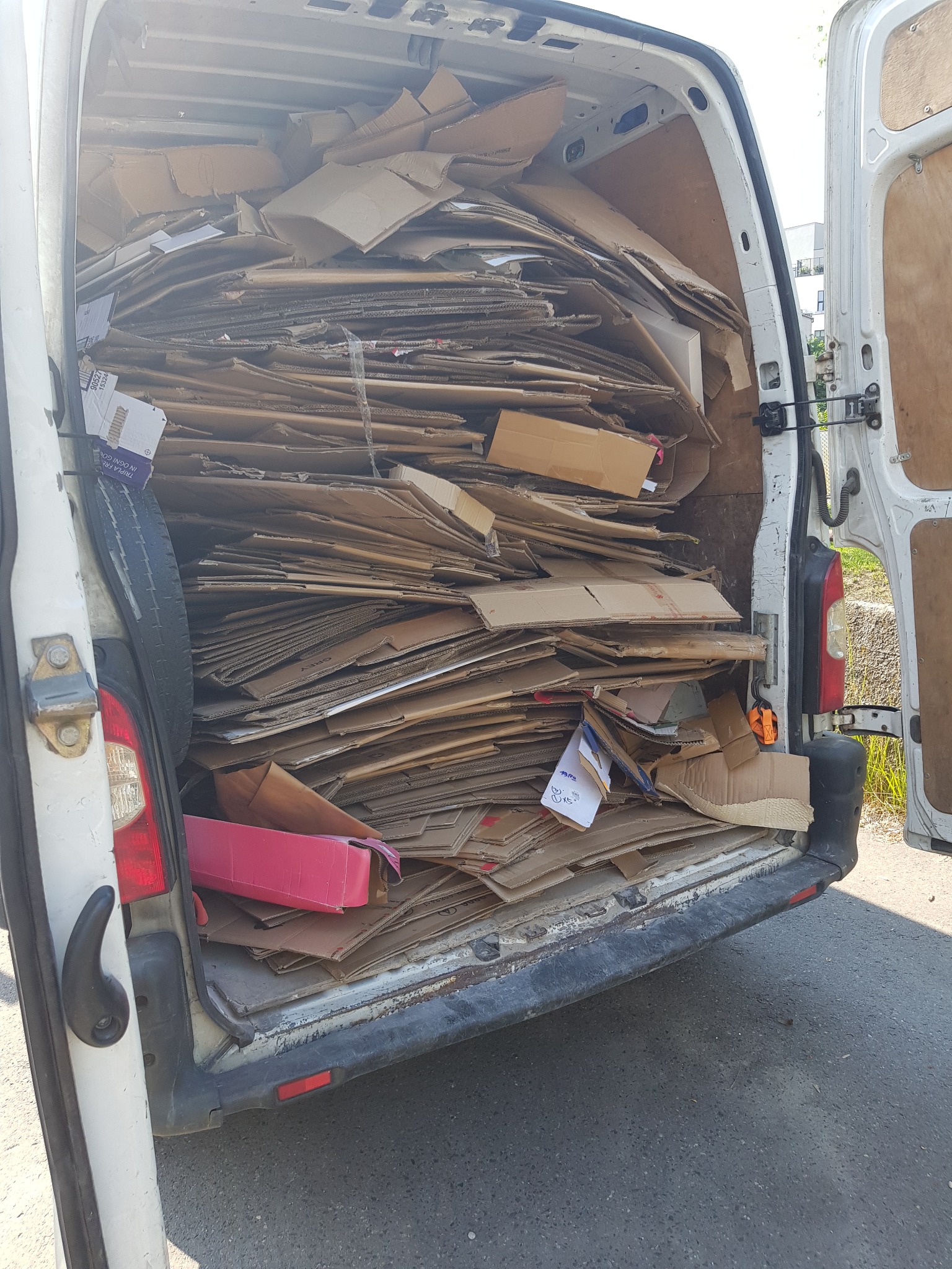 Wnętrze białego busa wypełnione po brzegi pogniecionymi kartonami, widoczne otwarte tylne drzwi i opona zapasowa po lewej stronie.